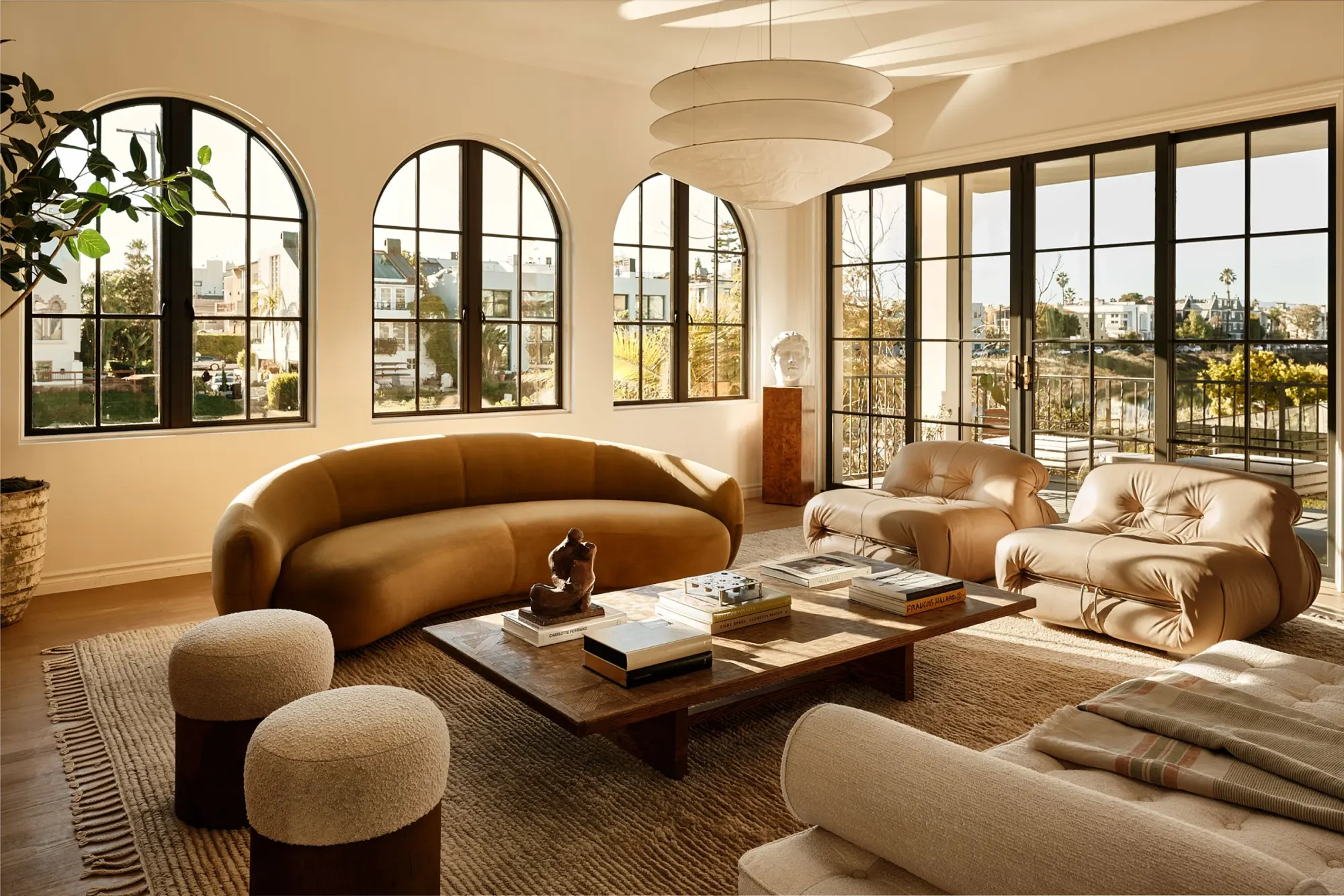 Chic living room with arched windows and warm light. Features brown curved sofa, plush armchairs, wooden table with books, and textured stools.