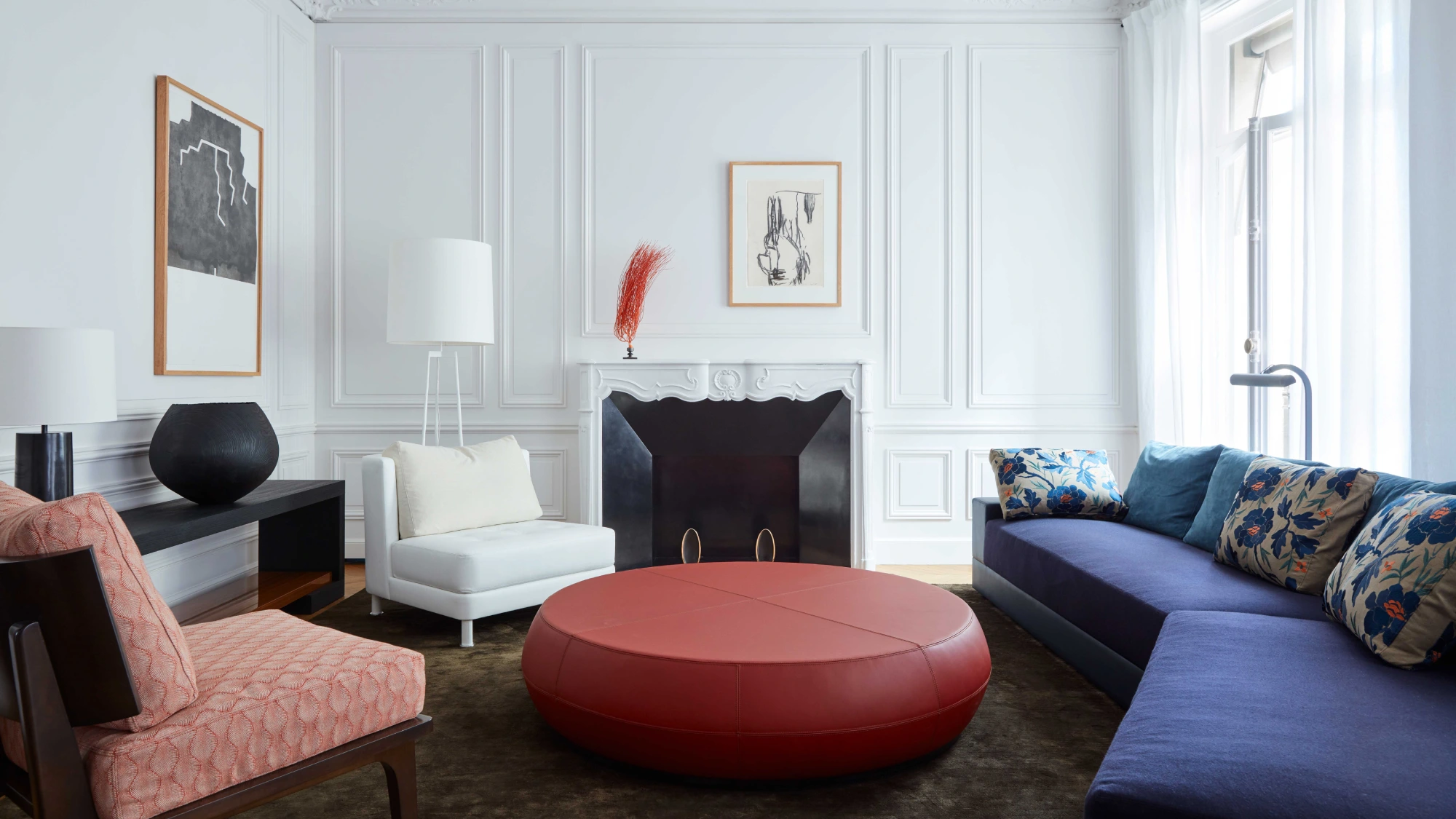 A modern living room with white paneled walls, a red circular ottoman, blue sofa with floral cushions, and a white armchair by a tall lamp. Cozy ambiance.