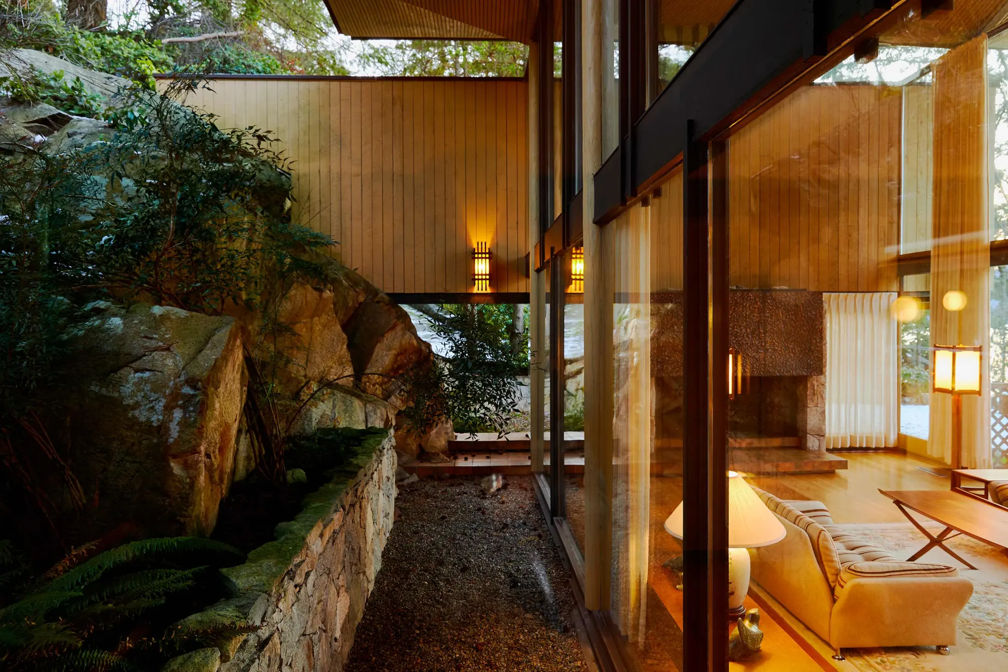 Warmly lit modern living room with floor-to-ceiling windows, a soft sofa, and a lamp. Outside, large rocks and greenery create a natural ambiance.