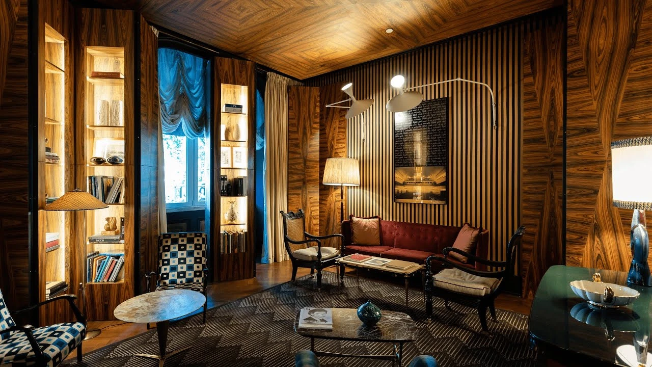 A cozy living room at Hotel Ververde in Lisbon with warm wooden walls, built-in shelves, elegant lighting, a burgundy sofa, and vintage armchairs.