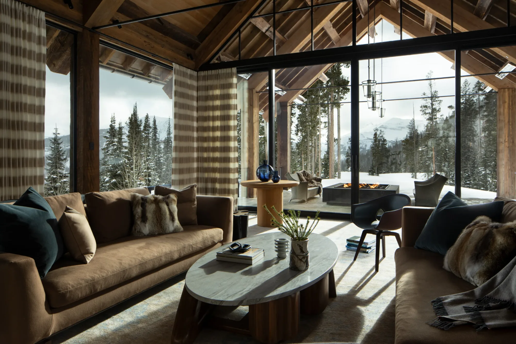 Cozy living room with brown sofas, fur pillows, and a marble coffee table overlooking snowy mountains through large glass doors, exuding warmth and luxury.