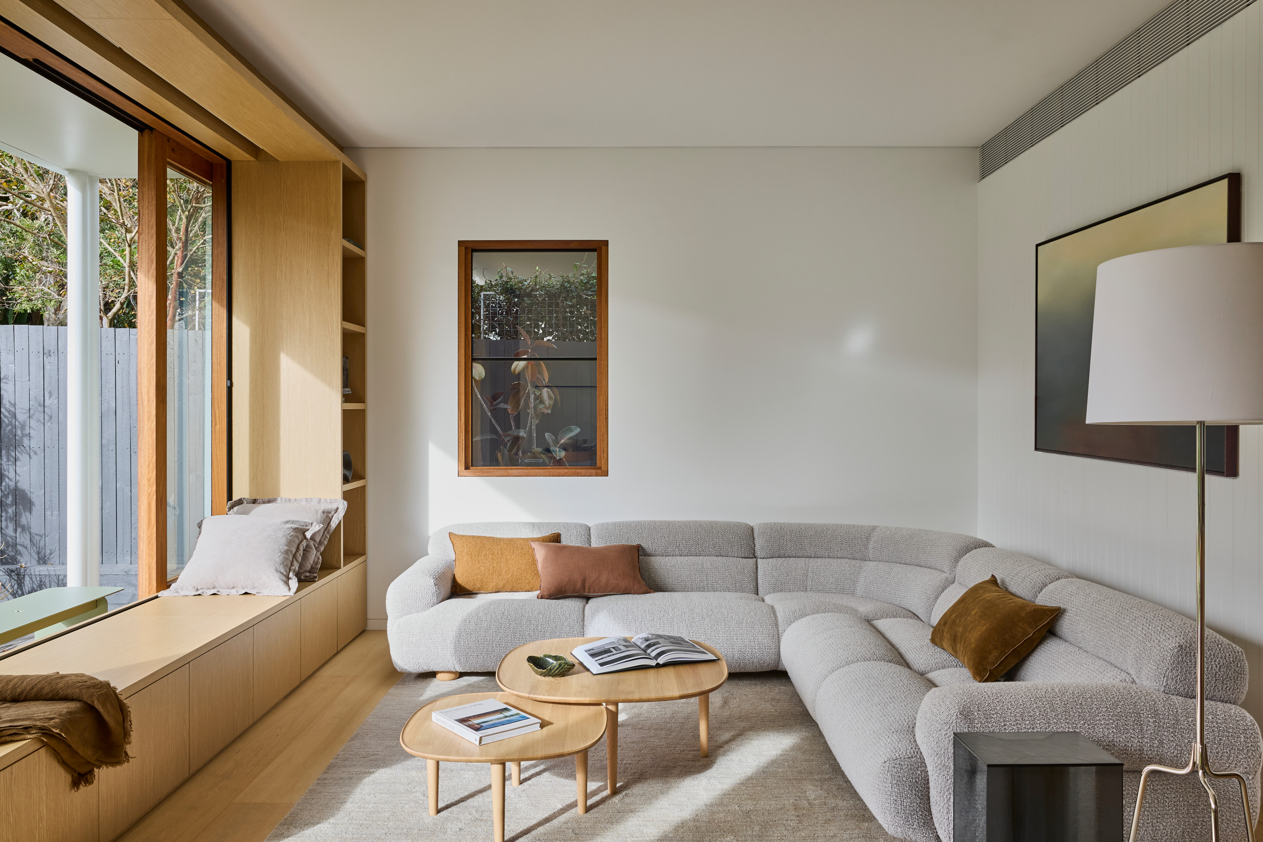 Modern living room with a light gray sectional sofa, colorful cushions, wooden tables, and large windows. Warm, cozy atmosphere with natural light, ideal for luxury interior design.
