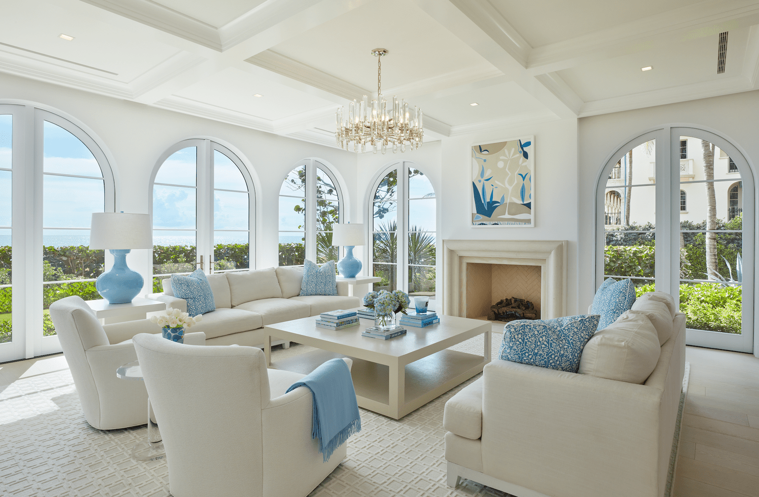 Bright, elegant living room with white sofas, blue cushions, and a central coffee table. Arched windows showcase a garden view. A chandelier adds sophistication.