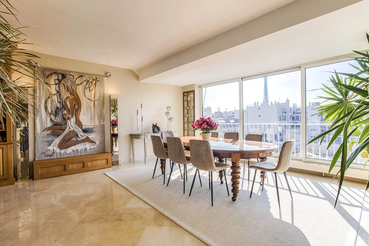 Bright dining room with a large wooden table and gray chairs, adorned by a vase of pink flowers. Floor-to-ceiling windows reveal a cityscape, including a distant Eiffel tower. Art features prominently on a wall, and lush plants add a warm, inviting feel.