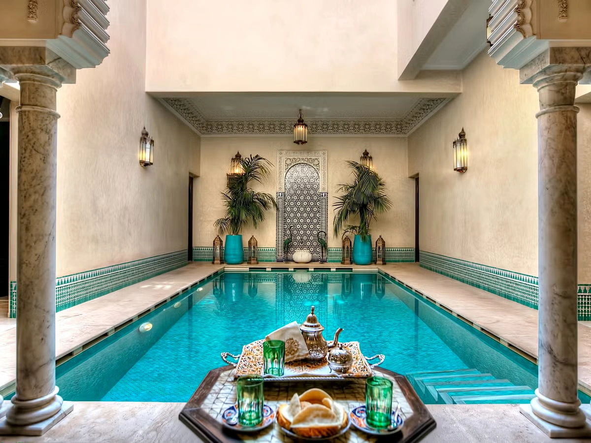 Luxurious Moroccan courtyard with a central turquoise pool, flanked by ornate columns. Foreground has a tray with tea set, evoking a tranquil ambiance.