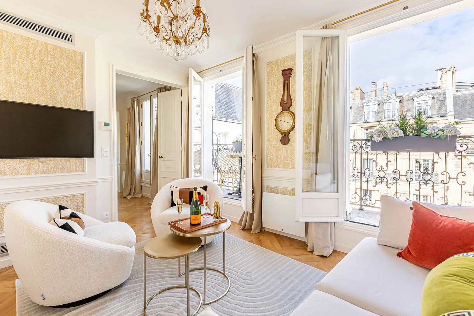Elegant living room with a chandelier, open French windows, and city view. White chairs, sofa with red and green pillows, and a wall-mounted TV. Warm, inviting atmosphere.