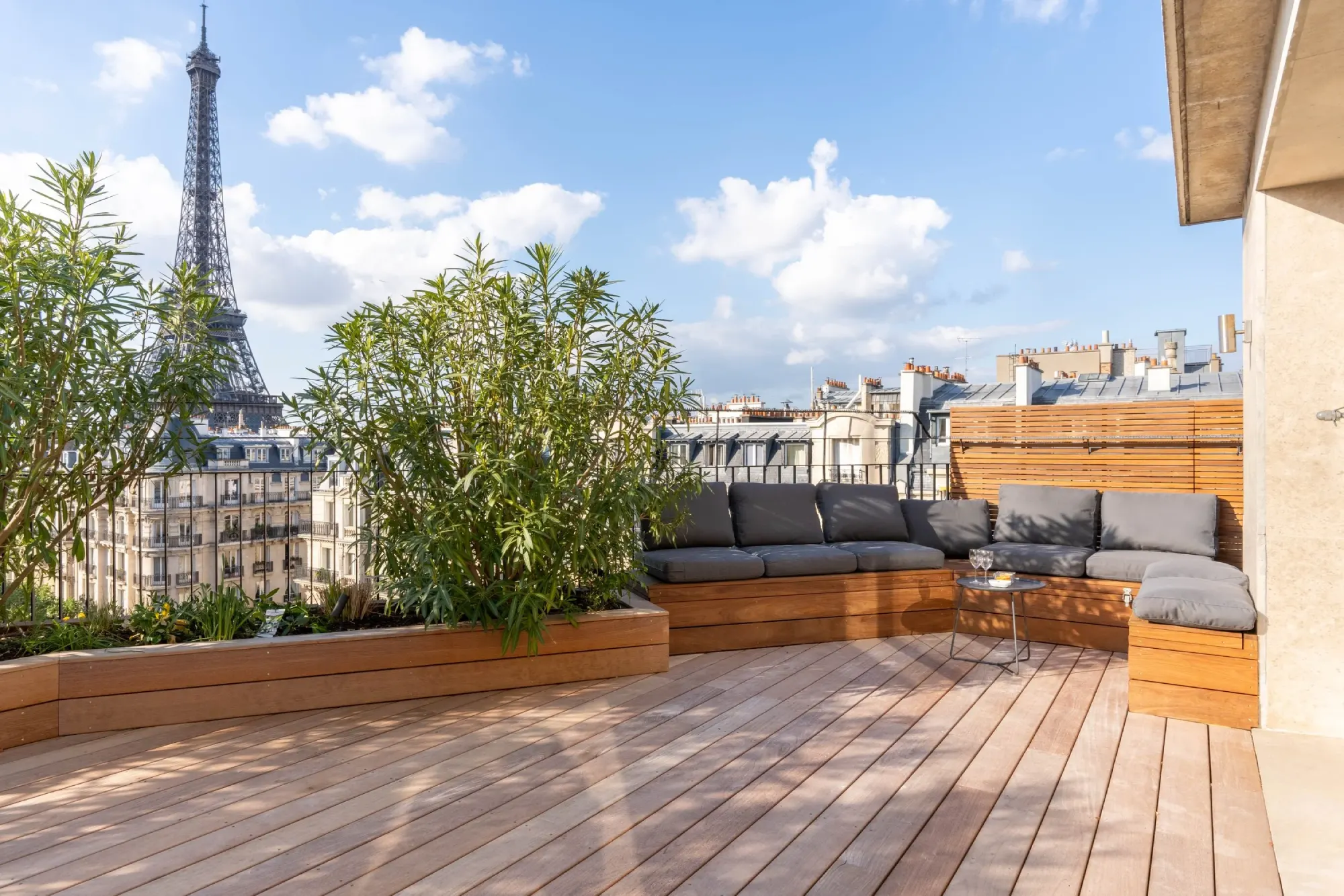 Rooftop terrace with wooden decking, gray cushioned seating, and lush plants. Eiffel Tower in the background against a blue sky with clouds.