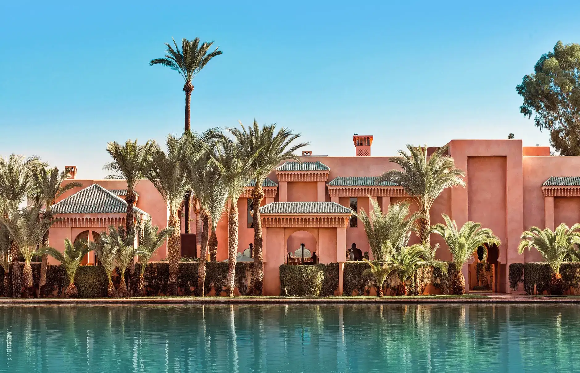 Luxurious villa with pink walls and green-tiled roofs, surrounded by palm trees, reflects in a tranquil blue pool under a clear sky.