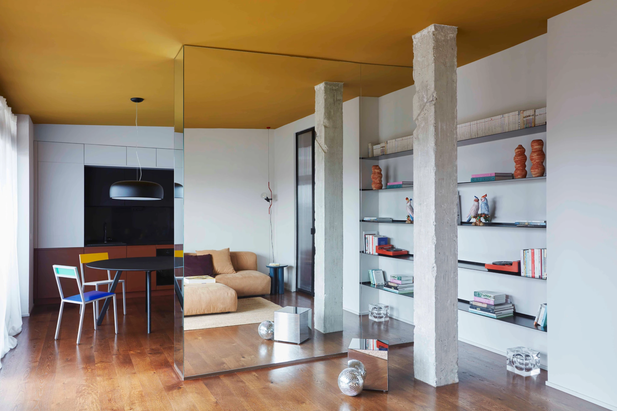 Modern living room with mustard ceiling, mirrored wall, and wooden flooring. Features a beige sofa, colorful chairs, shelves with books, and abstract decor.