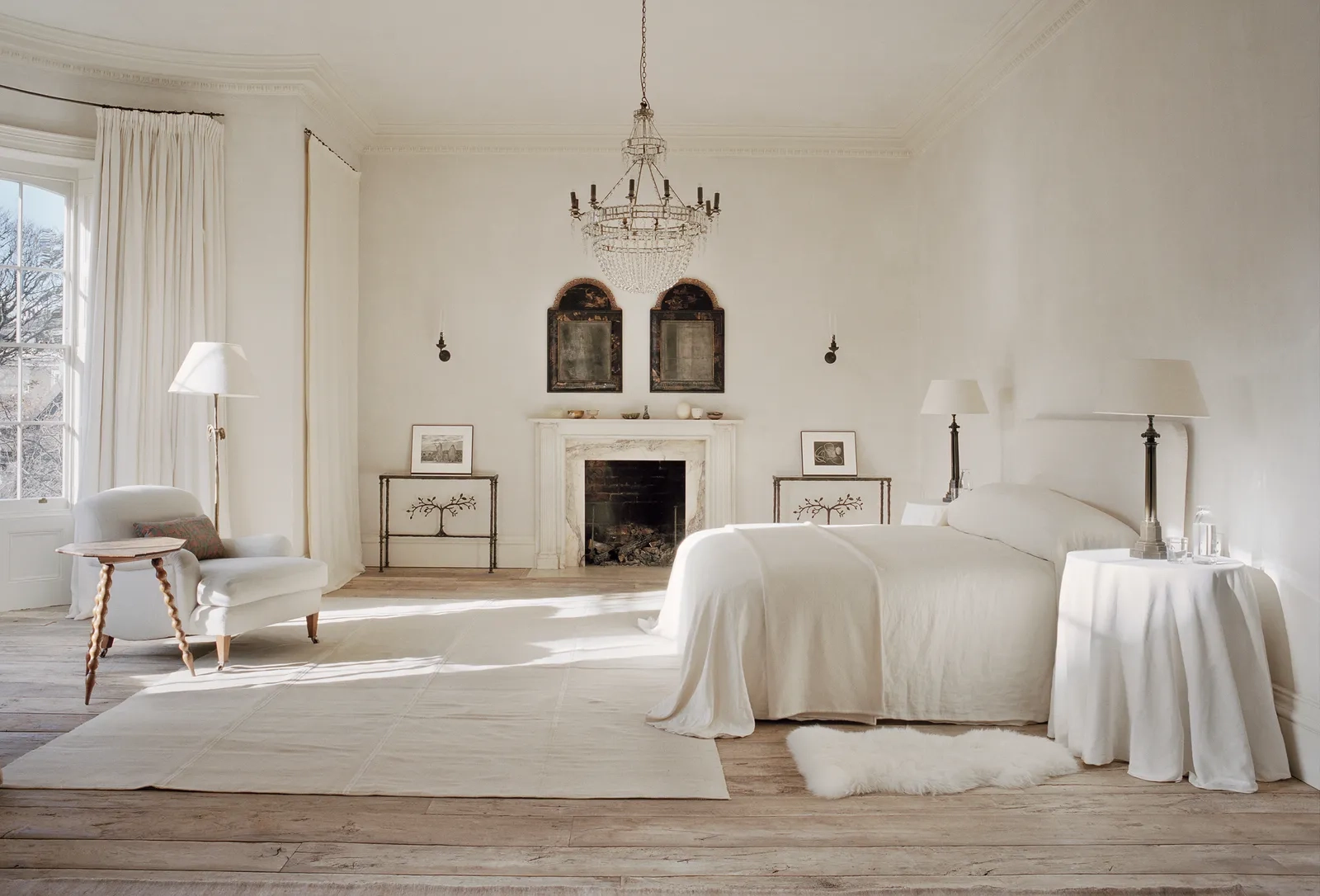 A serene, elegant bedroom with a white color palette features a plush bed, vintage chandelier, large windows, soft lighting, and a cozy fireplace.