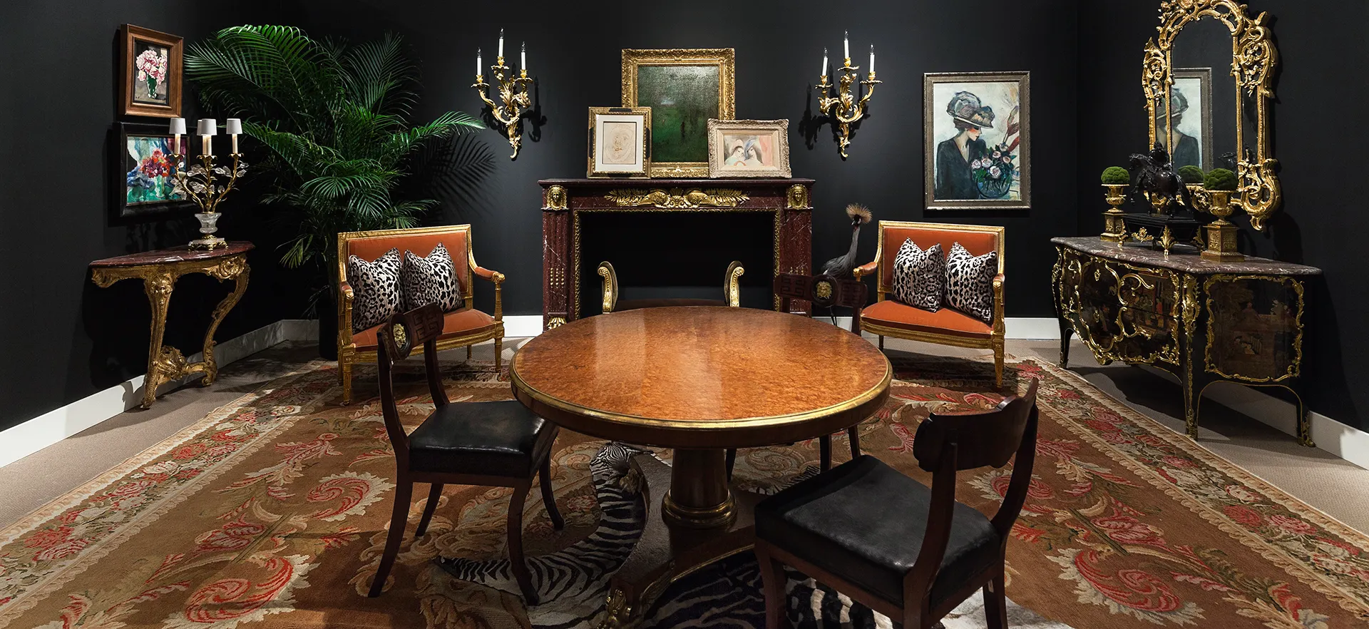 Luxurious room at Sotheby's, designed by Ryan Korban with elegant wooden furniture, including a round table and chairs. Opulent decor features gold-framed art, a mirror, and a fireplace.