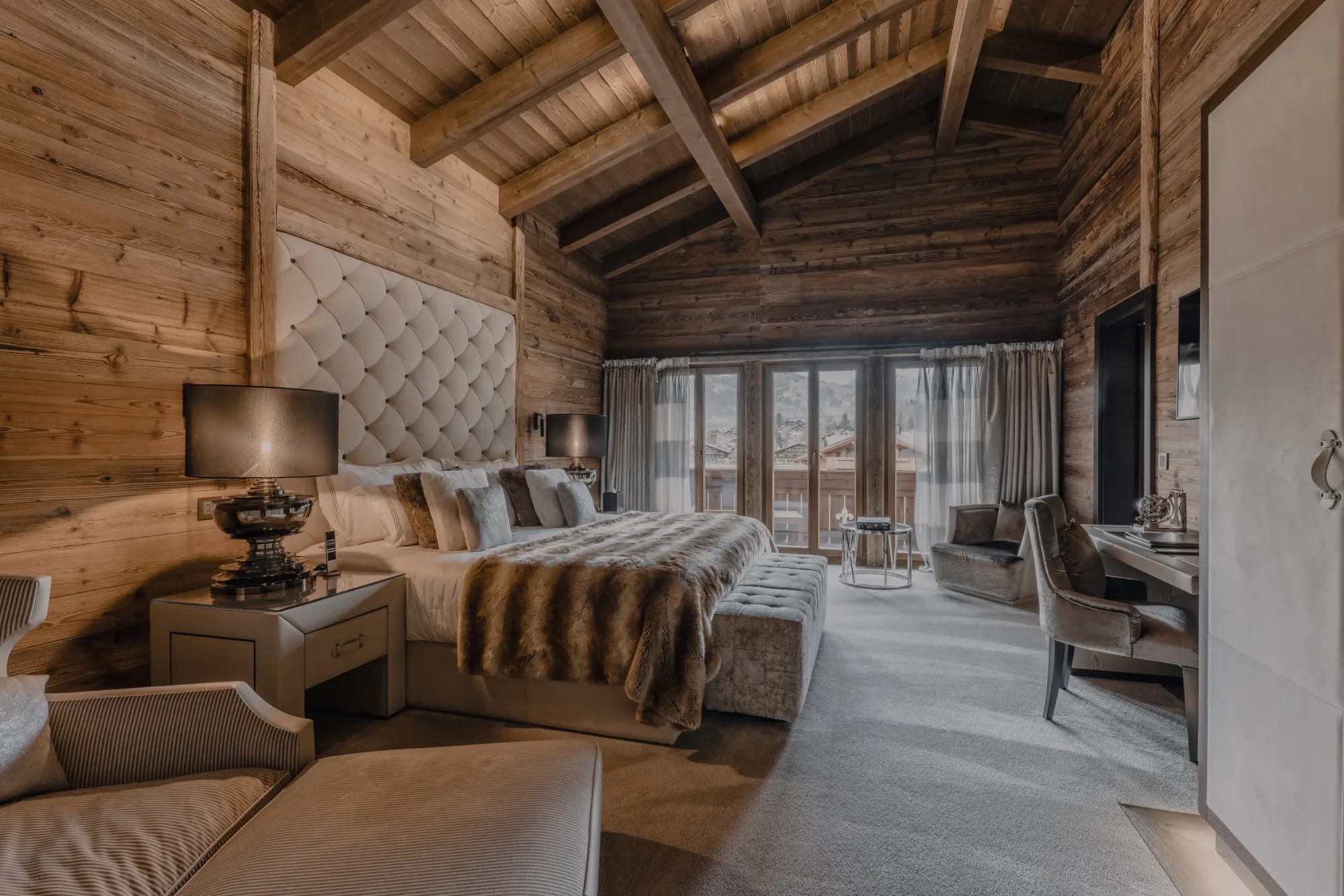 Luxurious wood-paneled bedroom with vaulted ceiling, a plush bed adorned with fur throw, elegant lamps, a cozy chair, and glass doors opening to a scenic view in the Ultima Gstaad hotel.