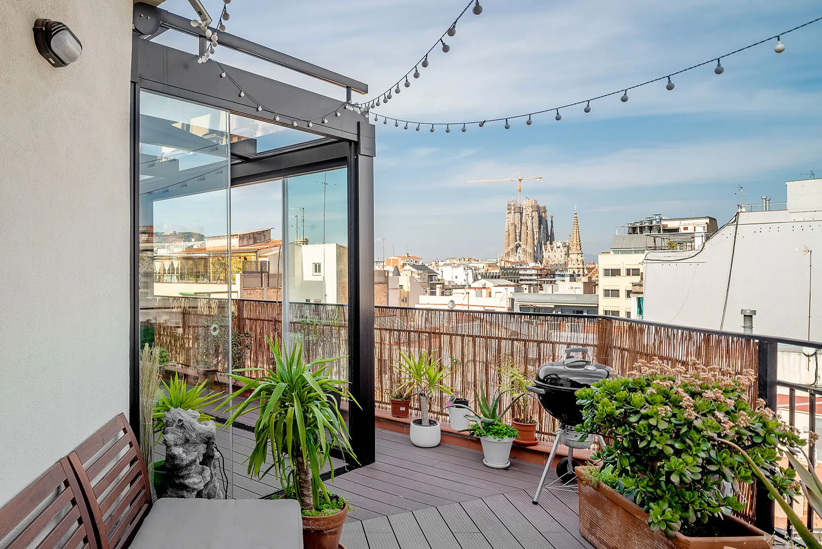 Rooftop terrace with wooden decking, potted plants, and string lights. A distant cathedral towers above city rooftops under a clear blue sky.