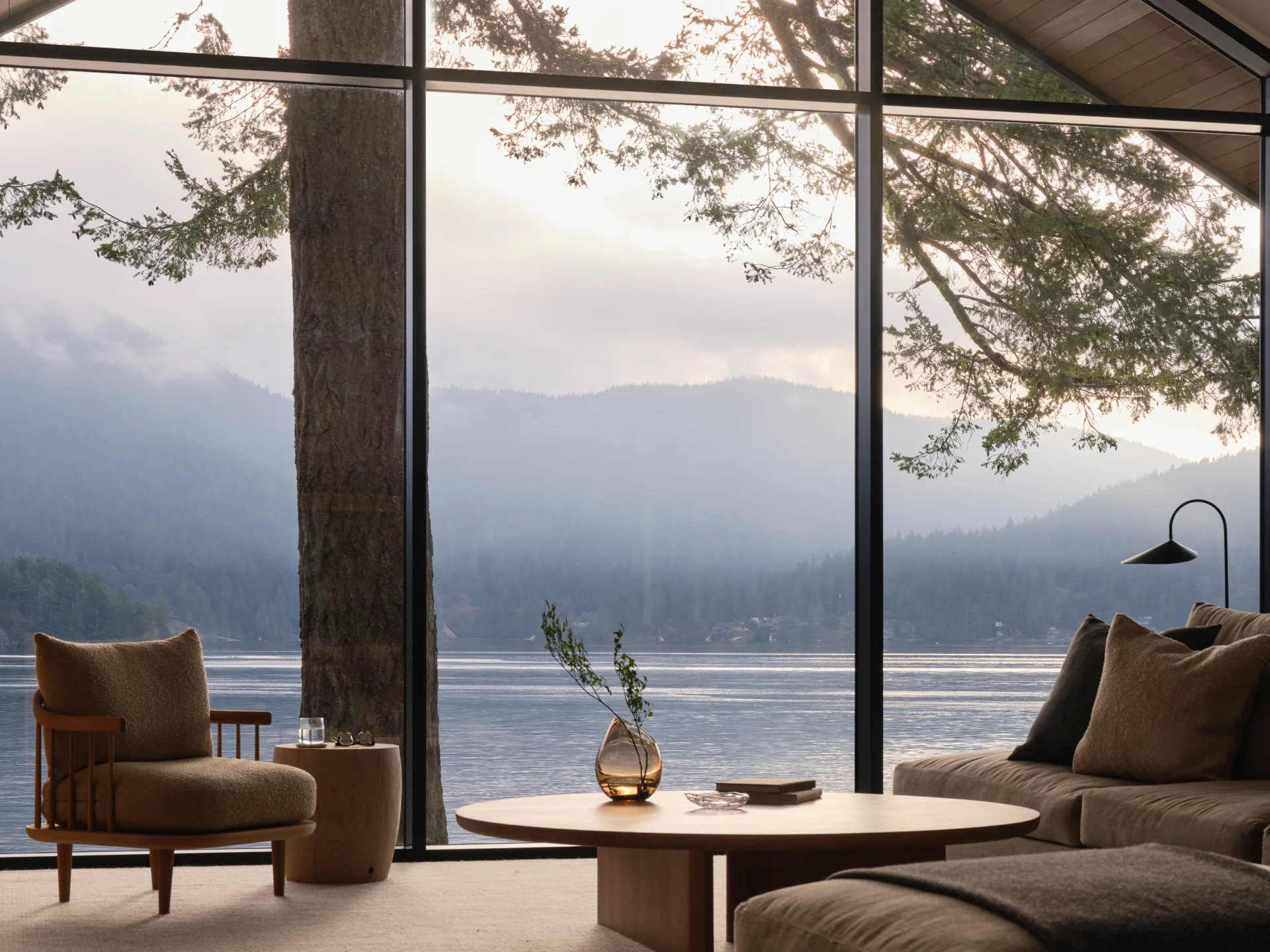 Spacious living room with large windows showcasing a serene lake and misty mountain view. Cozy armchair, round table, and elegant decor create a tranquil ambiance found in Vancouver Homes.
