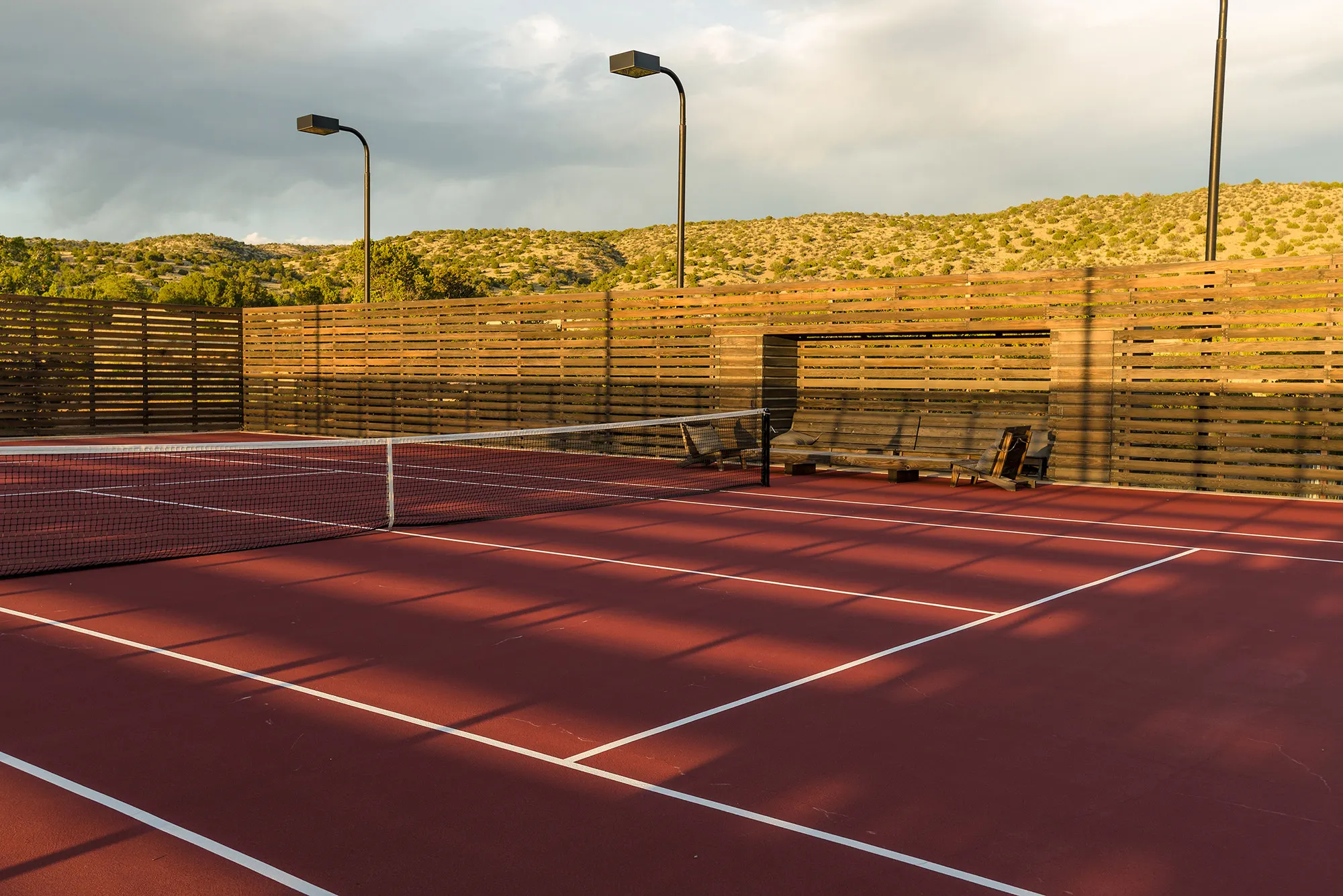 Sunlit tennis court inside Cerro Pelon Ranch with red surface, surrounded by wooden fencing. Hills and shrubs are visible in the background under a partly cloudy sky.