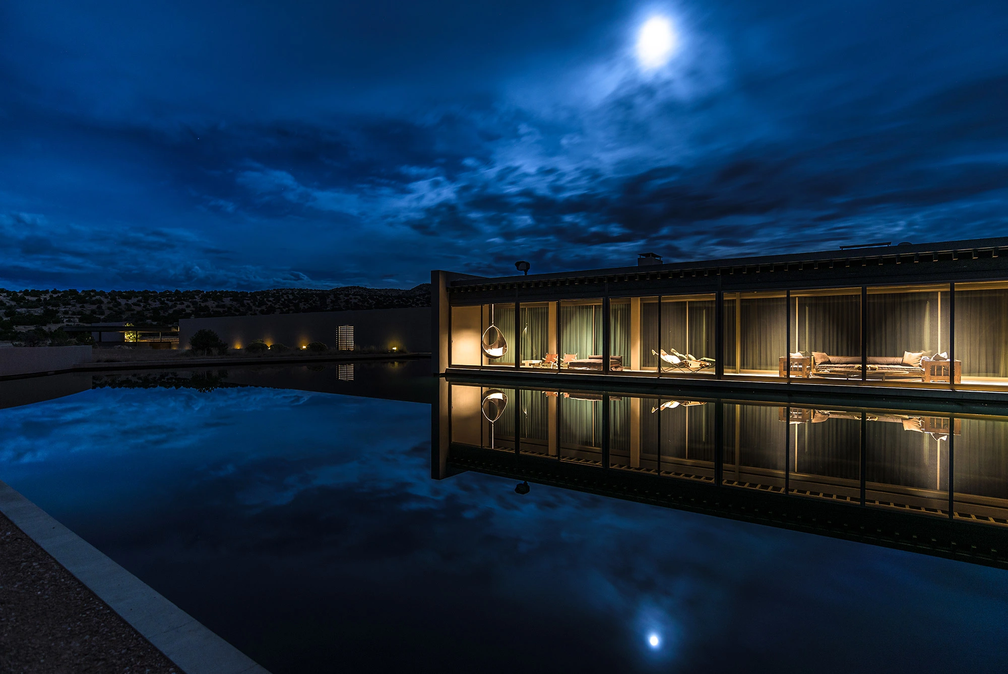 Modern building at Cerro Pelon Ranch with large windows is warmly lit at night. It reflects on a calm pool beneath a cloudy, moonlit sky, creating a serene ambiance.