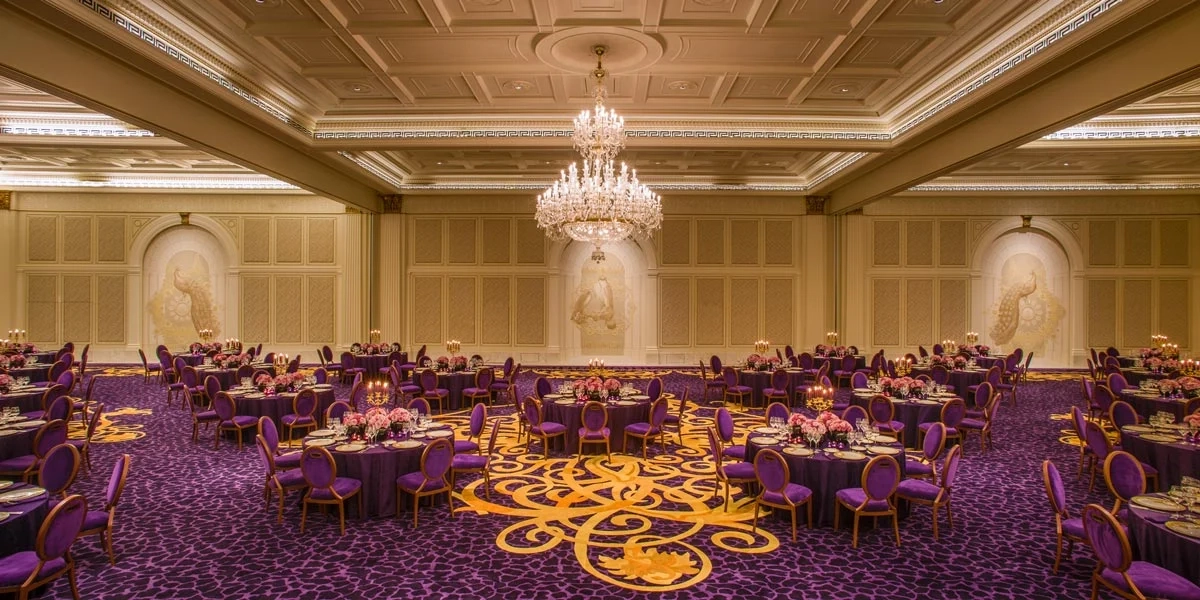 Luxurious gala ballroom at the Palazzo Versace Dubai with ornate chandeliers, purple and gold carpet, round tables with purple chairs, elegantly set for an event. 