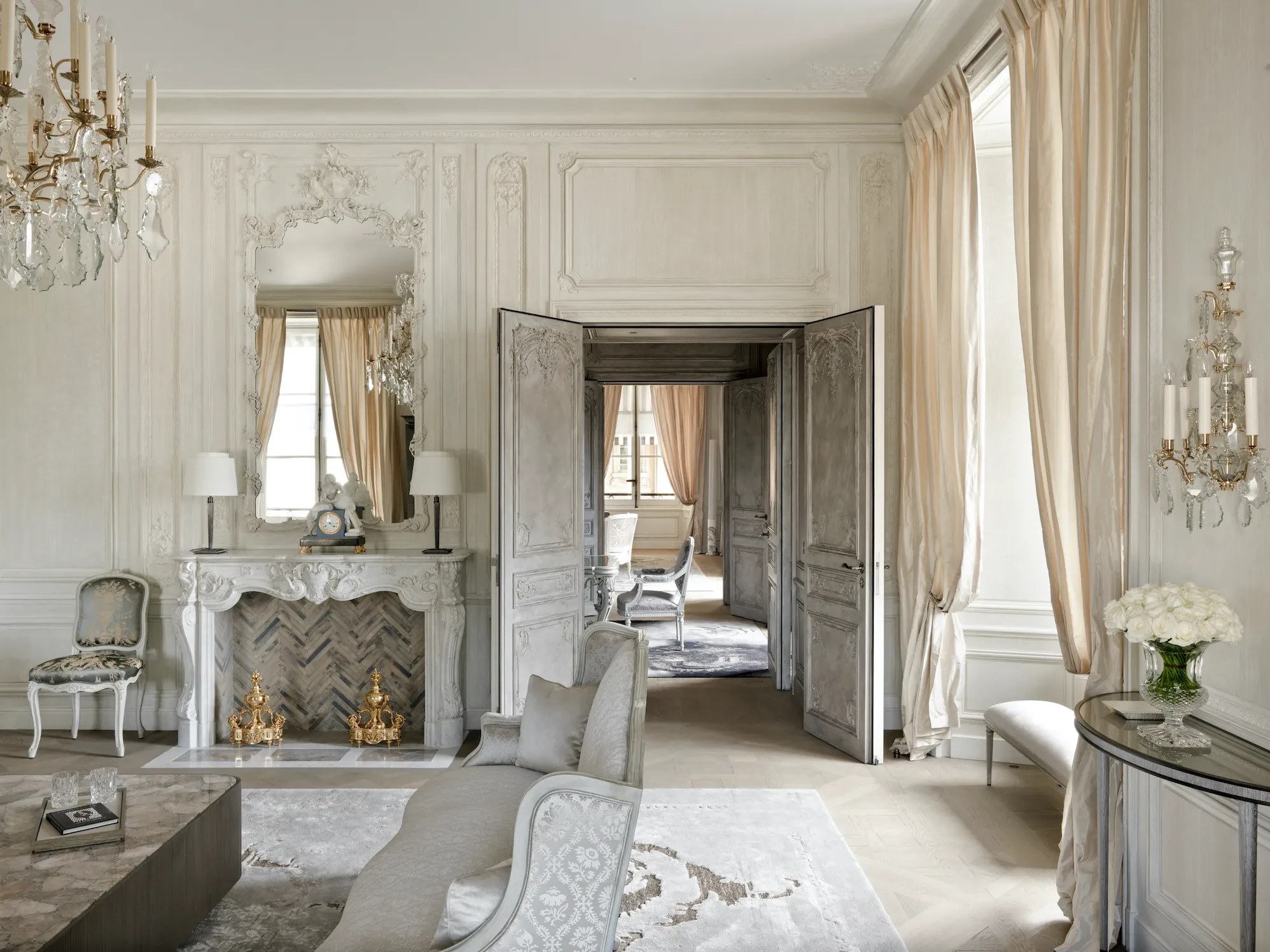 Elegant living room inside le Crillon hotel in Paris with ornate white walls, vintage fireplace, and chandeliers. 