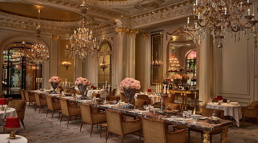 Luxurious dining room at the Plaza Athénée hotel, in Paris, with ornate chandeliers, elegant marble tables, and plush chairs. The room is adorned with gold accents and soft lighting, exuding opulence.