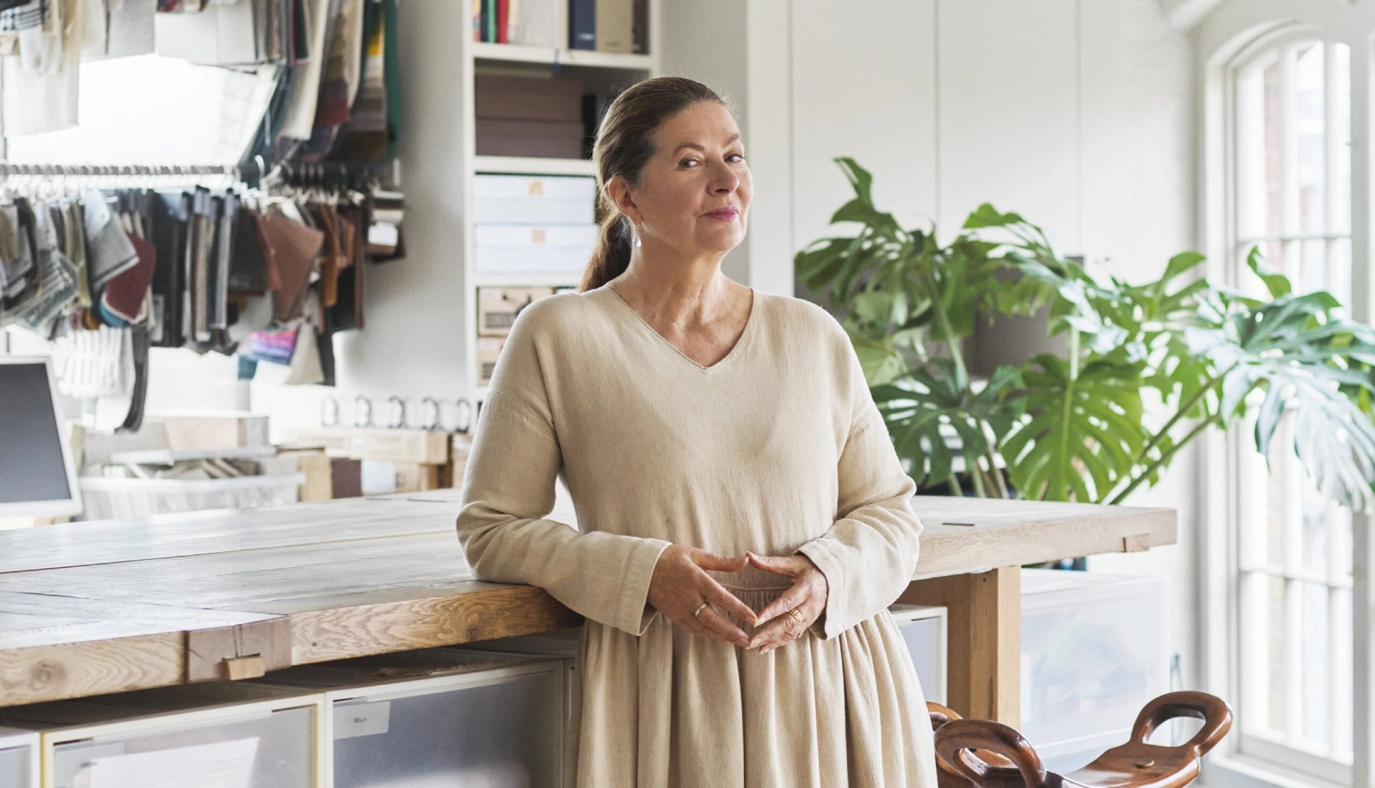 Interior designer Ilse Crawford in a beige dress stands confidently in a bright, organized workspace. Fabric samples and large plants are visible, creating a creative, peaceful vibe.