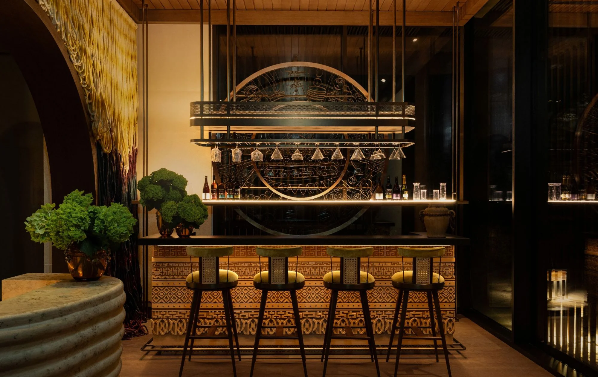 Elegant, dimly lit bar at the Intercontinental Red Sea Resort, decorated by Kristina Zanic with five stylish stools, ornate decorative patterns, hanging wine glasses, lush green plants, and warm, inviting ambiance.