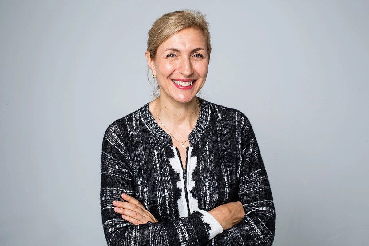 Interior designer Kristina Zadic, wearing a black and white patterned cardigan, stands against a plain gray background, arms crossed confidently.