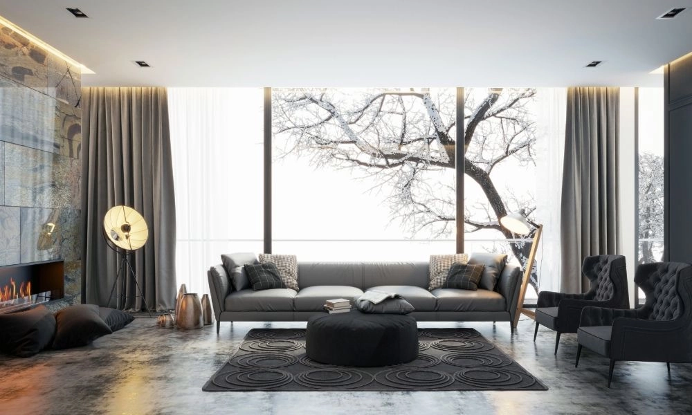 Modern living room with large windows showcasing a winter landscape. Gray sofa, armchairs, and round ottoman create a cozy, elegant ambiance.