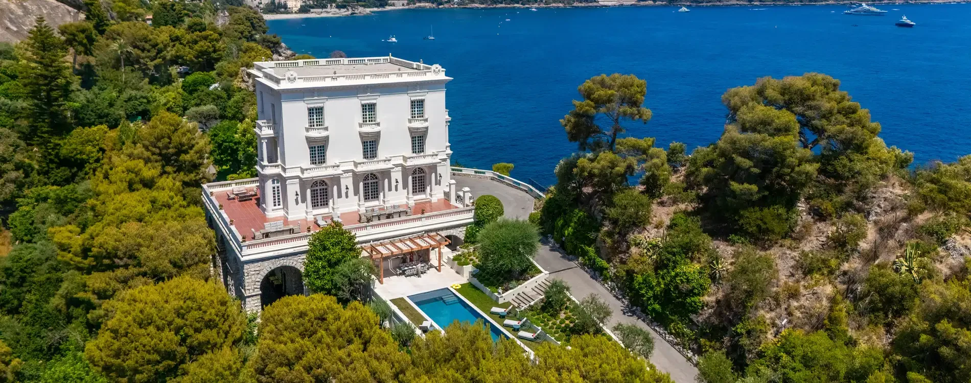 Aerial view of a luxurious white villa in Monaco surrounded by lush greenery, overlooking a vibrant blue sea. The scene conveys elegance and tranquility.