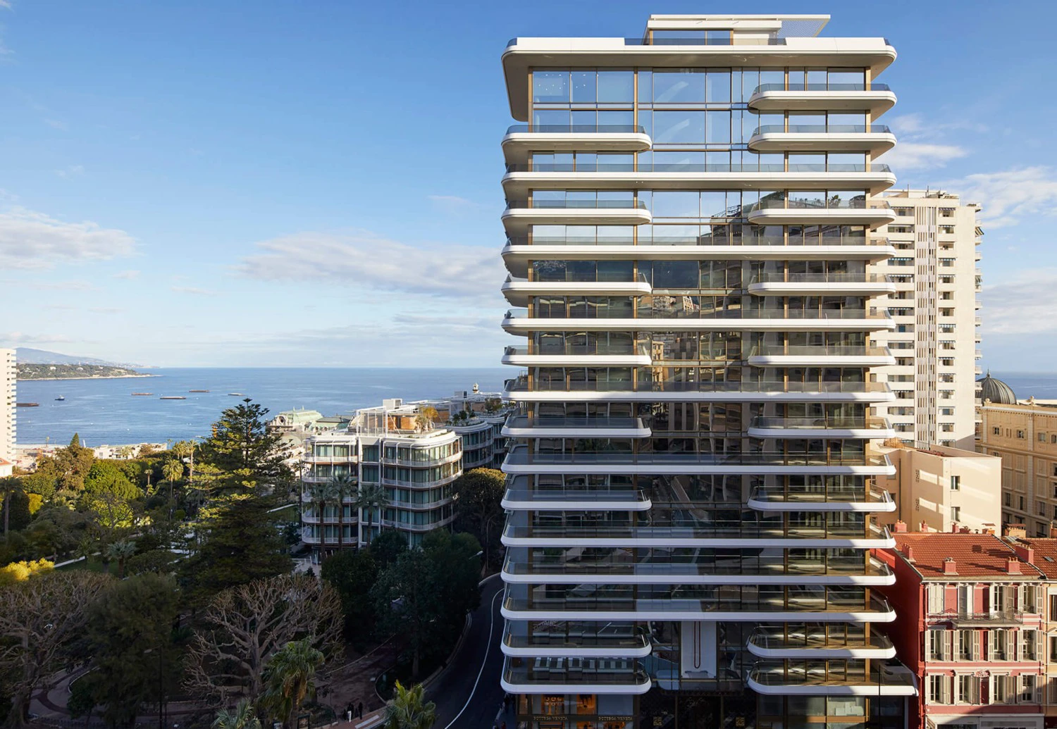 Modern high-rise building in Monaco with glass facade and balconies against a coastal backdrop. Surrounding trees and ocean in the distance under a clear blue sky.