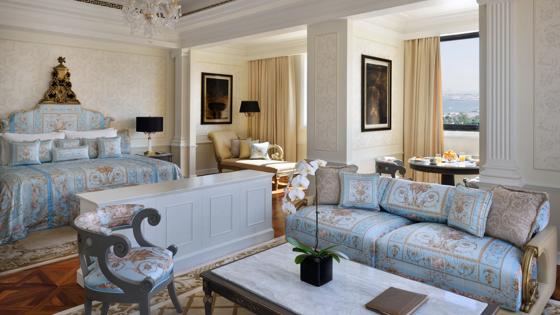 Luxurious Imperial Suite at the Palazzo Versace Dubai with opulent blue and gold decor. Features a large bed, elegant sofa, chandelier, and a window with a city view. 