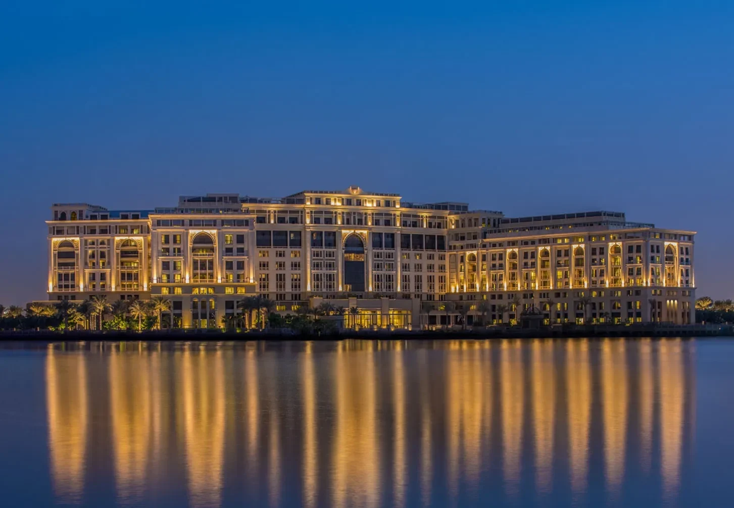 Palazzo Versace Dubai reflecting on a calm body of water at dusk. The building's warm lights create a serene and luxurious atmosphere against the deepening blue sky.