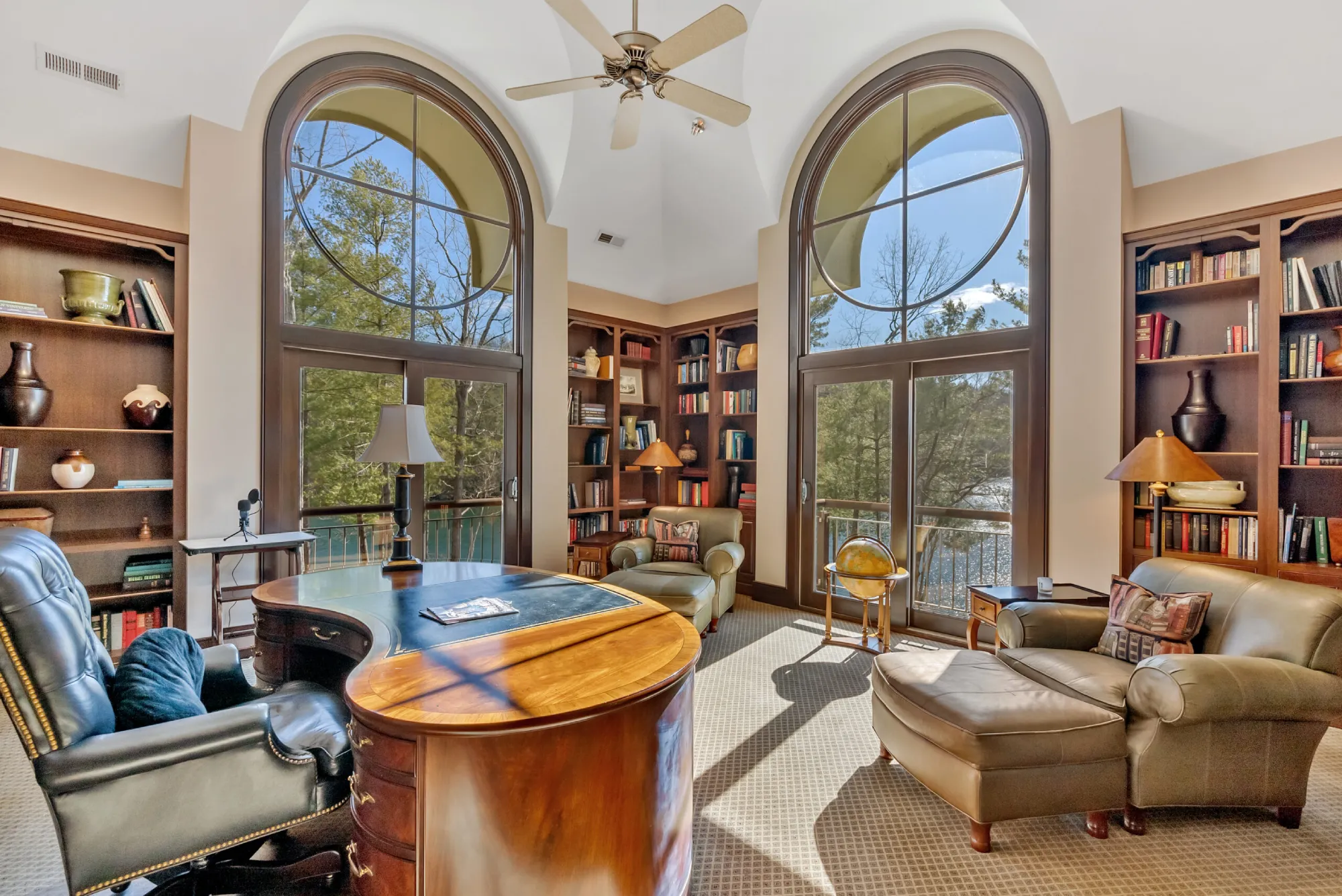 Elegant home reading nook with large arched windows, wooden shelves filled with books, leather armchairs, and a central wooden desk.
