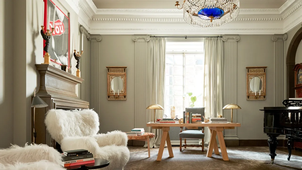 Elegant living room by Ilse Crawford with plush white chairs, a wooden desk, and a grand piano. Warm tones, a chandelier, and soft lighting create a cozy, sophisticated ambiance.