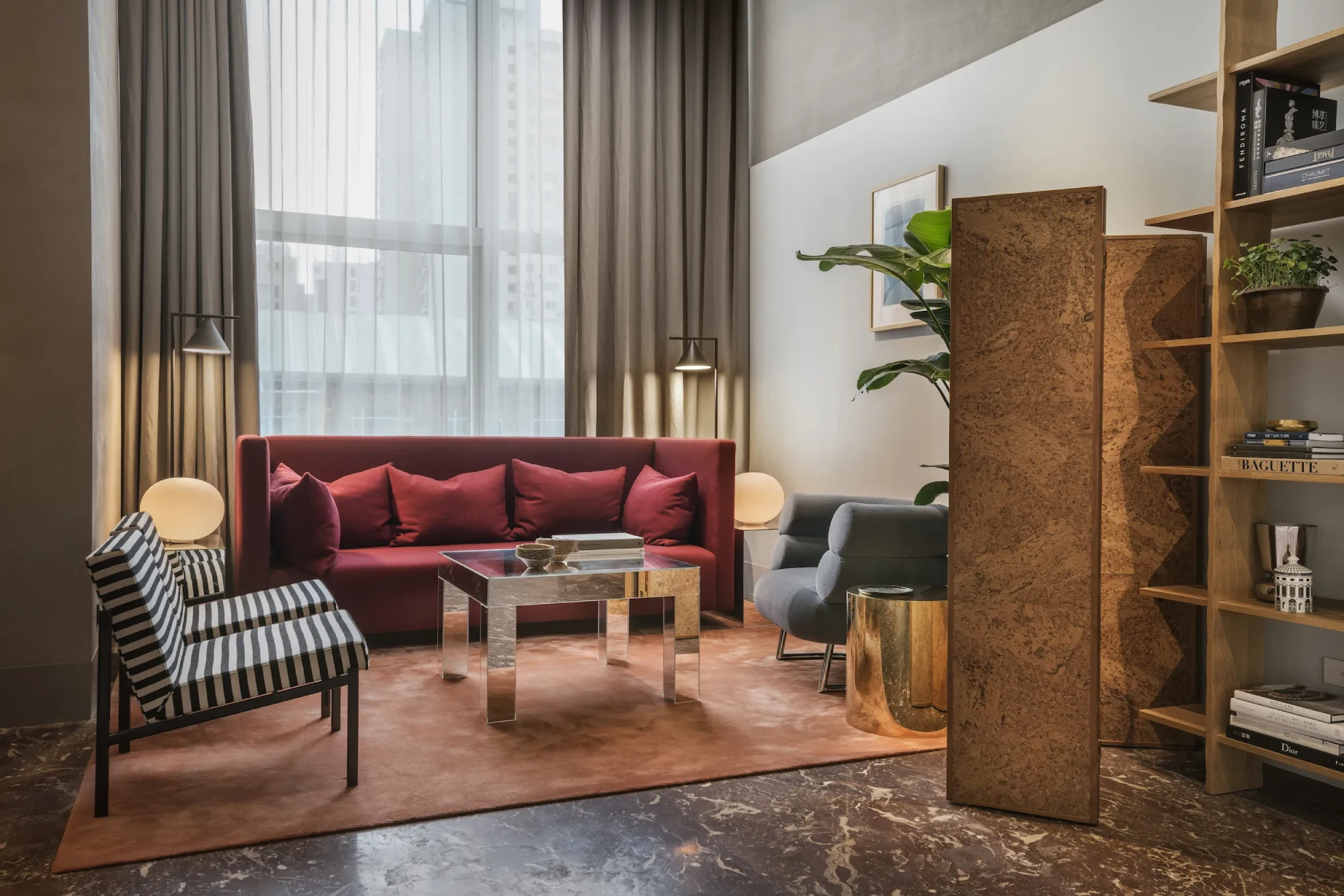 Modern living room by Studioilse by Ilse Crawford with a red sofa, striped chair, and glass coffee table on a rust rug. Tall window, beige curtains, and stylish decor create a cozy ambiance.