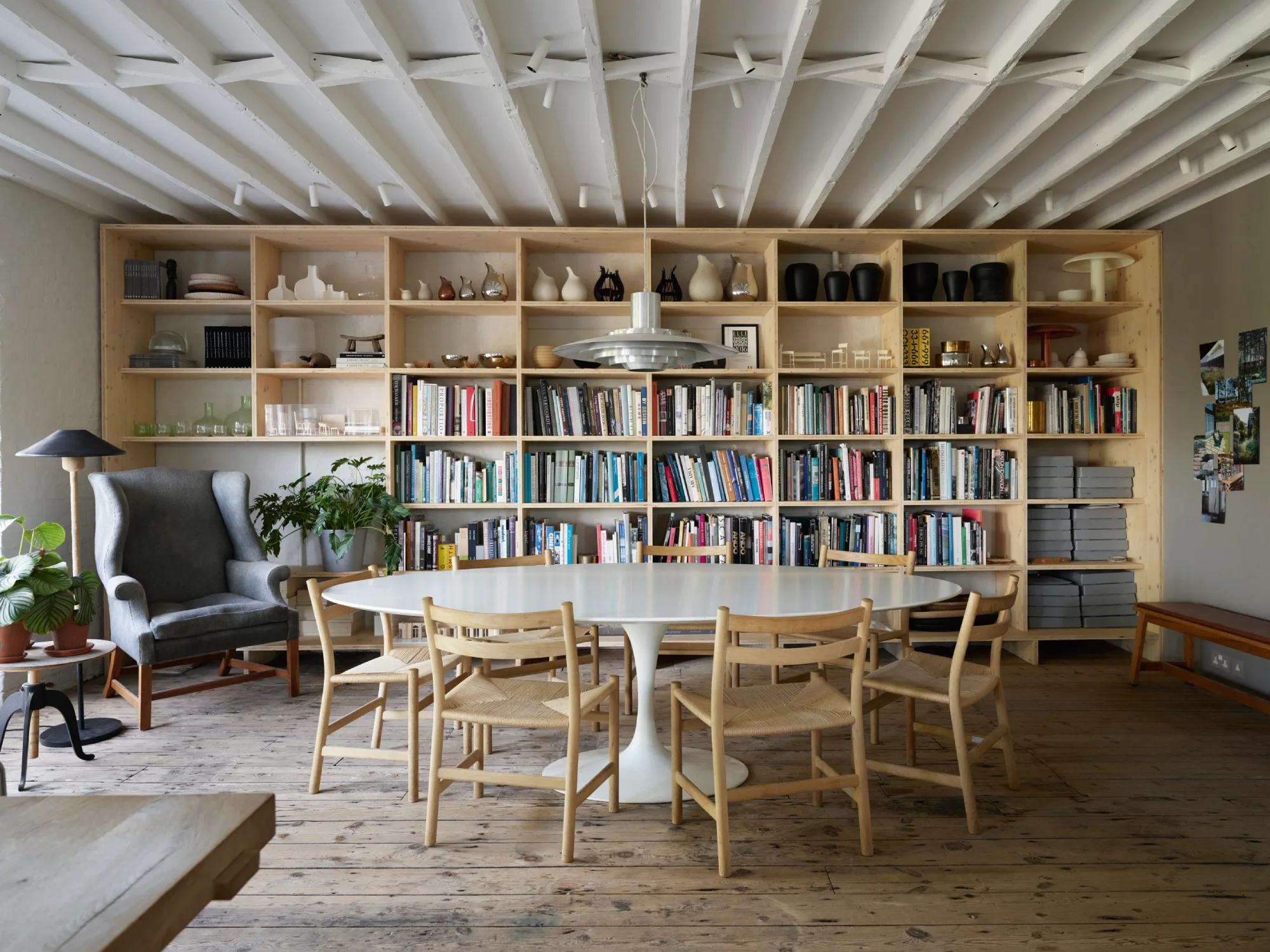 Stylish home library by Ilse Crawford with wooden shelves filled with books and decor. A white table, chairs, and a gray armchair create a cozy, modern reading space.