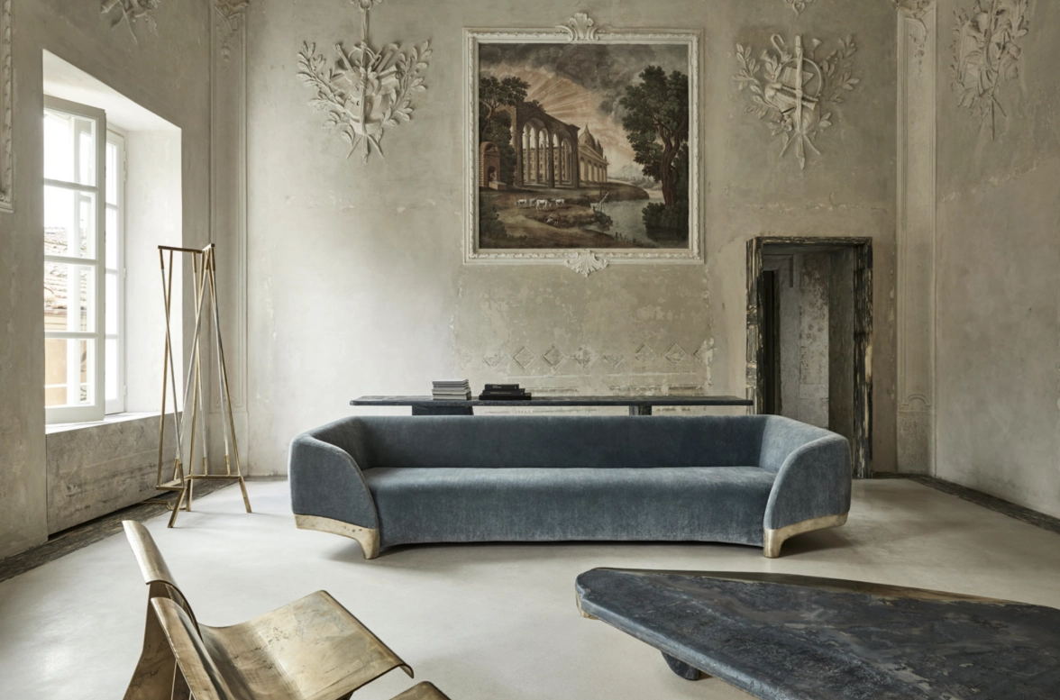 Elegant room at Palazzo Digerini by Vincenzo de Cotiis with a blue velvet sofa, rustic beige walls adorned with intricate molding, a large classical painting, and minimalistic wooden furniture.