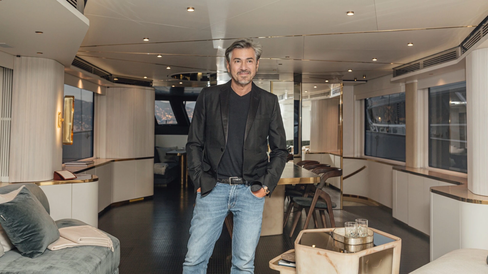 Vincenzo de Cotiis in a black blazer and jeans stands confidently in a modern, luxurious yacht interior. The setting is elegant, with soft lighting and sleek furnishings.
