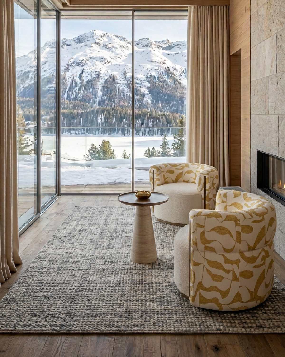 Two patterned armchairs and a side table by a fireplace, overlooking snow-covered mountains through floor-to-ceiling windows.